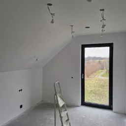 Poddasze w trakcie wykańczania: białe ściany, surowy beton na podłodze, drabina aluminiowa, okno z widokiem na krajobraz oraz niedokończone instalacje oświetleniowe zwisające z sufitu.