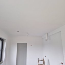 Krzysztof Krzyzowski. MACH-POL - Wnętrze świeżo wykończonego pokoju z białymi ścianami i sufitem; widoczna drewniana drabina, otwory pod oświetlenie i instalacje elektryczne, okno po lewej stronie.