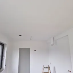 Wnętrze świeżo wykończonego pokoju z białymi ścianami i sufitem; widoczna drewniana drabina, otwory pod oświetlenie i instalacje elektryczne, okno po lewej stronie.