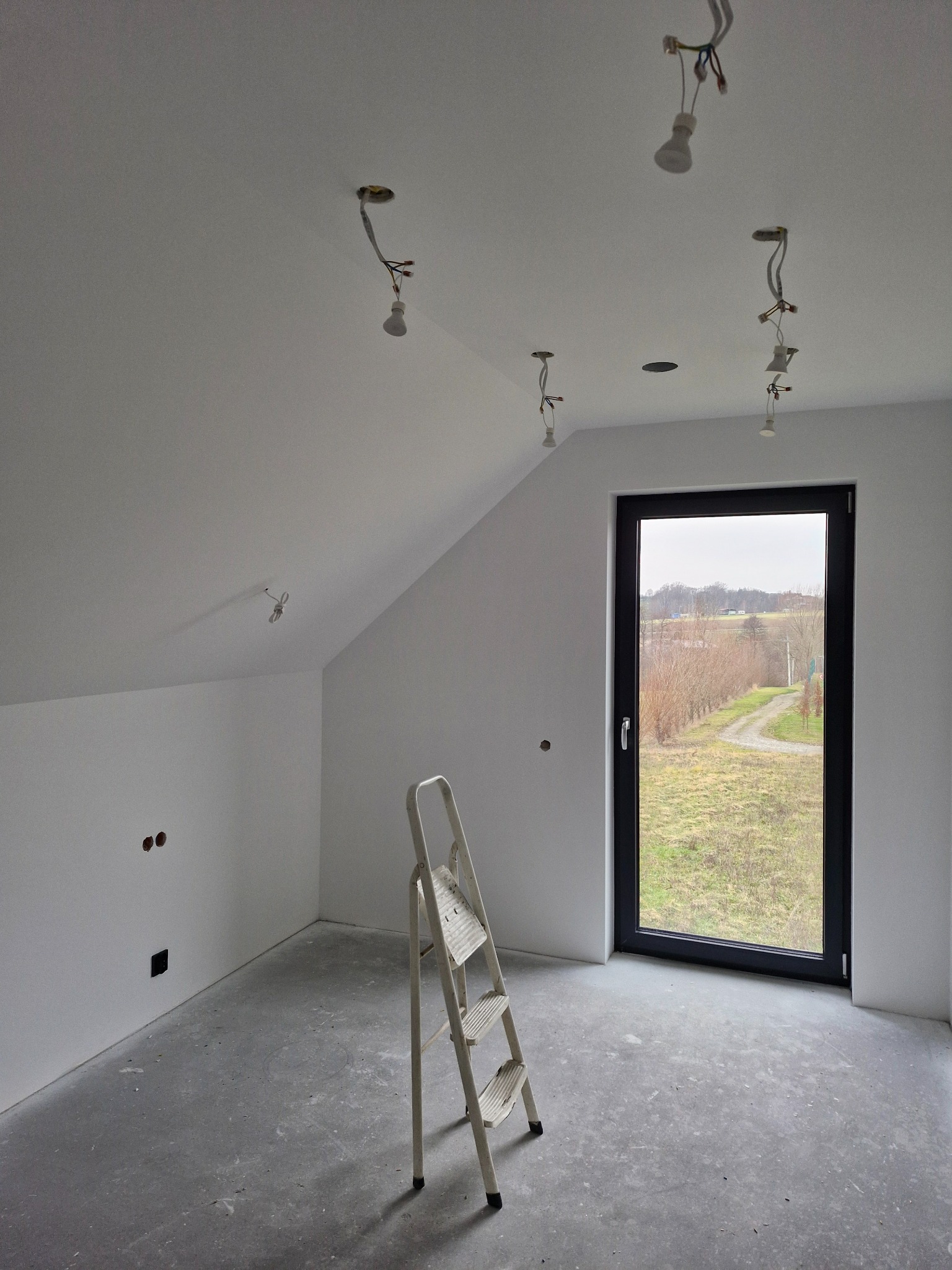 Poddasze w trakcie wykańczania: białe ściany, surowy beton na podłodze, drabina aluminiowa, okno z widokiem na krajobraz oraz niedokończone instalacje oświetleniowe zwisające z sufitu.
