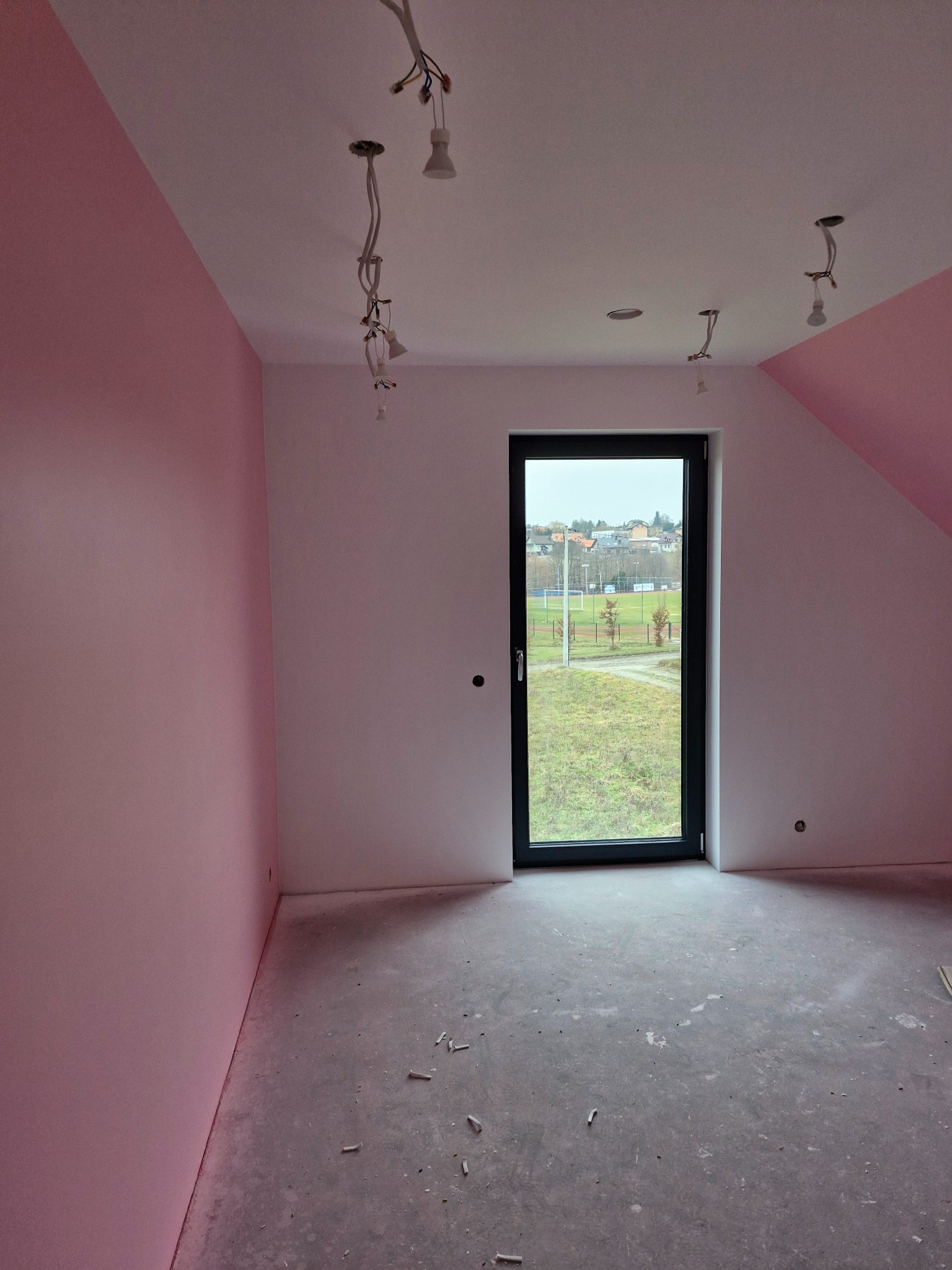 Pokój w trakcie prac wykończeniowych: ściany częściowo pomalowane na różowo, surowa podłoga, widoczne kable oświetleniowe i okno z widokiem na krajobraz.