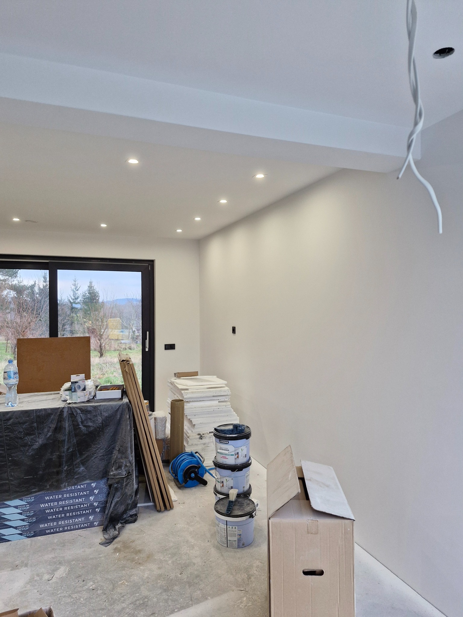 Wnętrze w trakcie malowania: ściany pomalowane na biało, widoczne puszki z farbą, przewody zwisające z sufitu, okno z widokiem na zewnątrz, podłoga zabezpieczona folią.