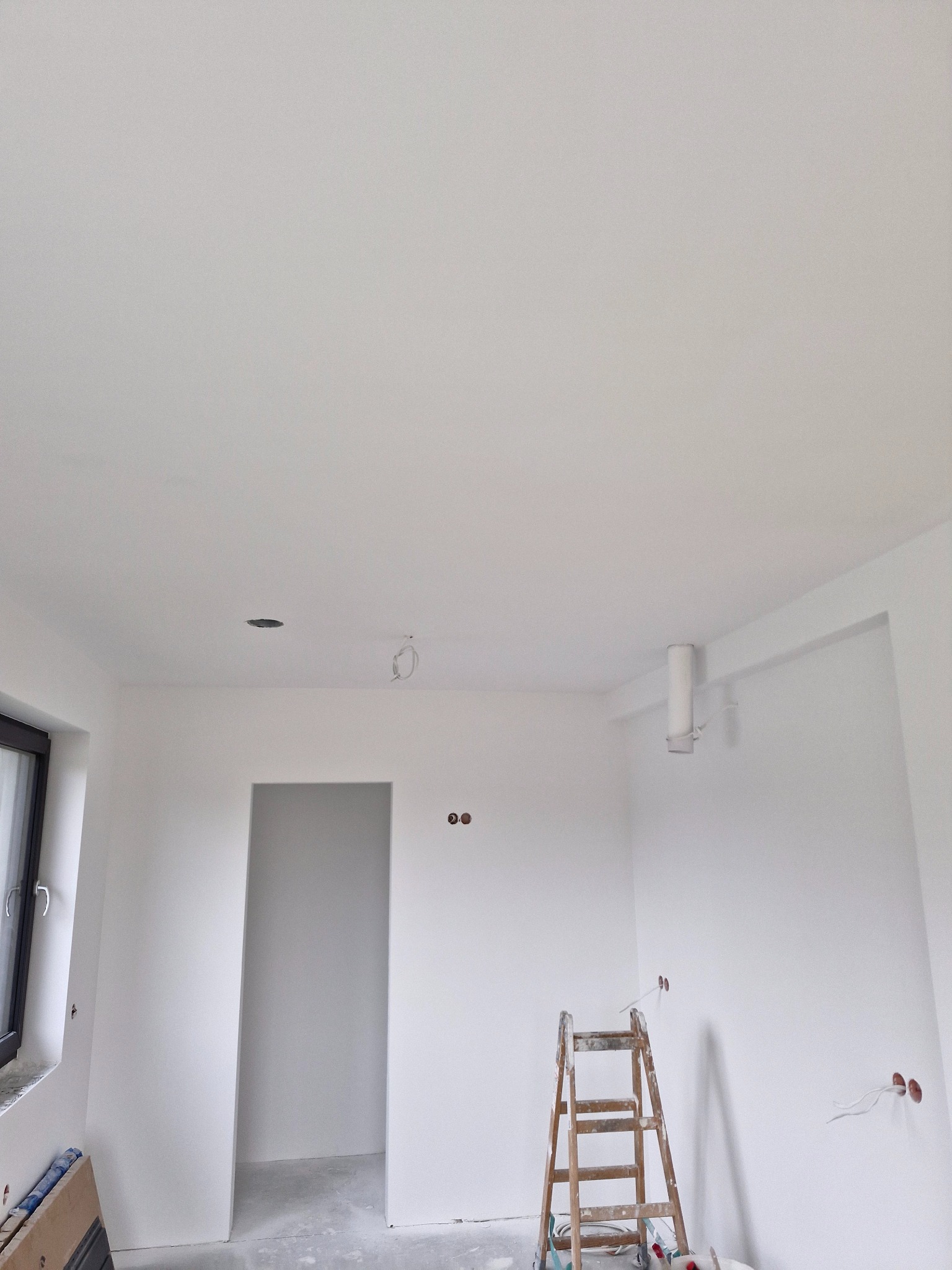 Wnętrze świeżo wykończonego pokoju z białymi ścianami i sufitem; widoczna drewniana drabina, otwory pod oświetlenie i instalacje elektryczne, okno po lewej stronie.