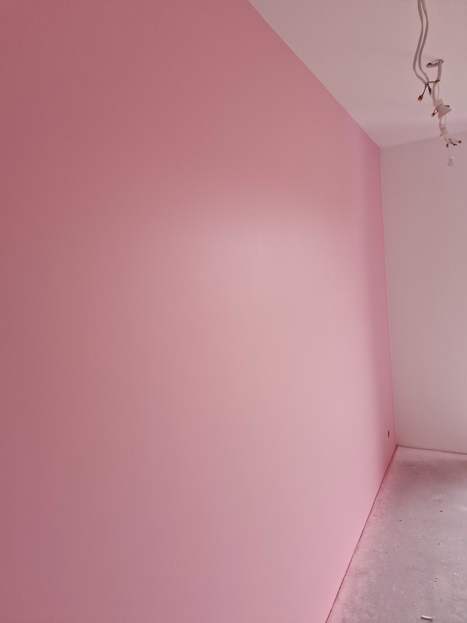 Różowa ściana w nowo wykończonym pokoju, widoczne przewody elektryczne wystające z sufitu, szary betonowy podkład podłogowy.