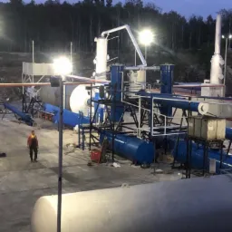 Instalacja do produkcji oleju opałowego z widocznym pracownikiem, zbiornikami i systemem rur w kolorach niebieskim i czerwonym, oświetlona lampami.