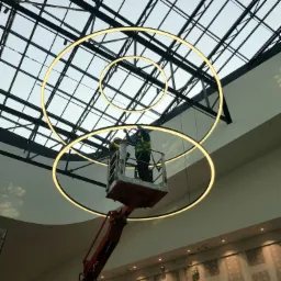 Dwóch elektryków w podnośniku koszowym montuje okrągłe lampy LED pod szklanym dachem w galerii handlowej.