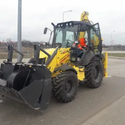 Żółta koparko-ładowarka B110B New Holland z operatorem w pomarańczowej kamizelce w kabinie, zaparkowana na kostce brukowej w pochmurny dzień.