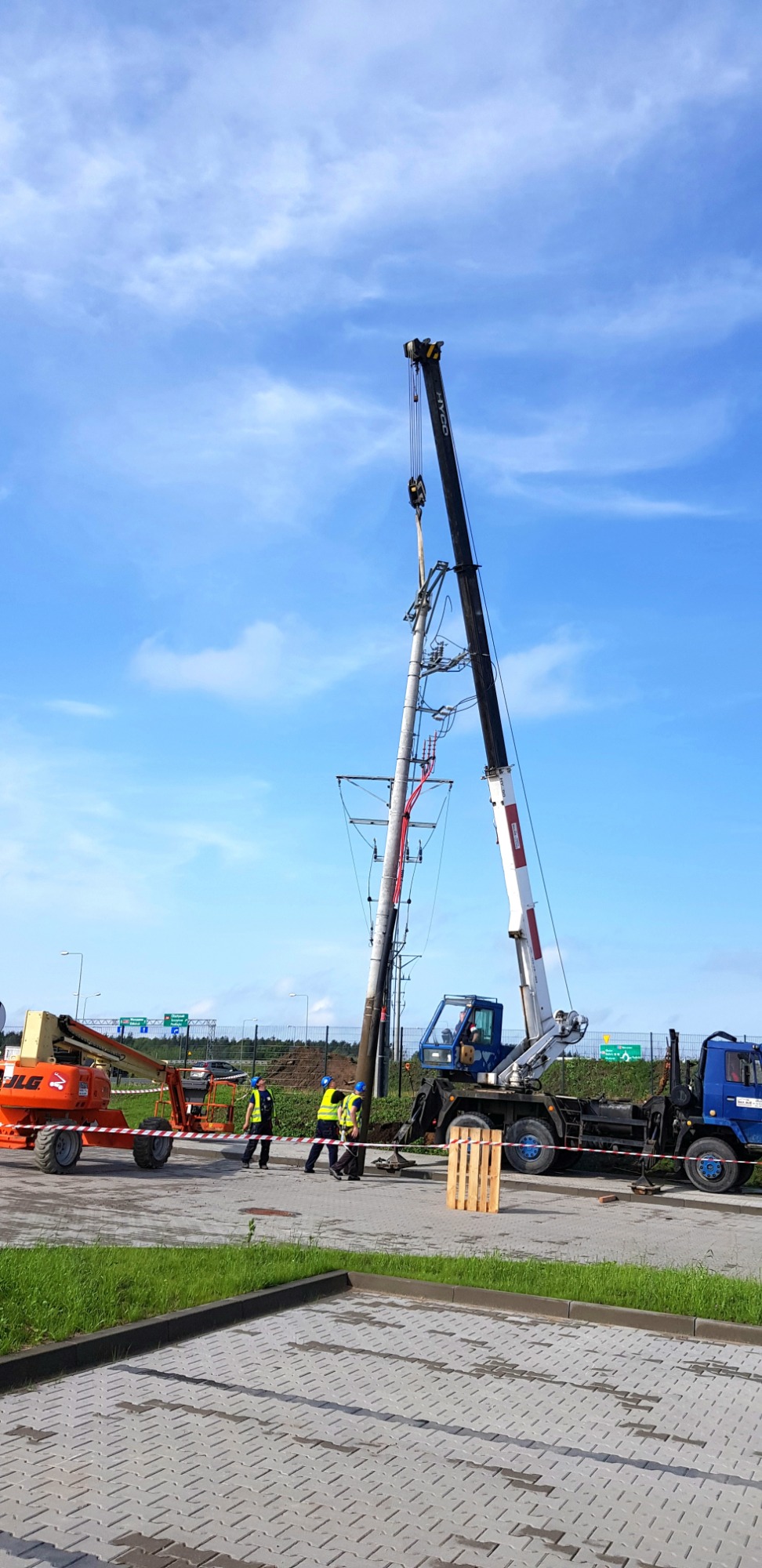 Montaż słupa energetycznego z użyciem dźwigu samochodowego, widoczny podnośnik koszowy i pracownicy w kaskach i kamizelkach odblaskowych, teren zabezpieczony taśmą.