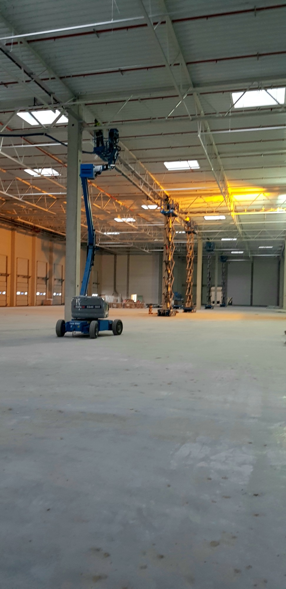 Wnętrze hali produkcyjnej z podnośnikami koszowymi podczas instalacji pod sufitem, widoczne elementy konstrukcyjne dachu i oświetlenie.