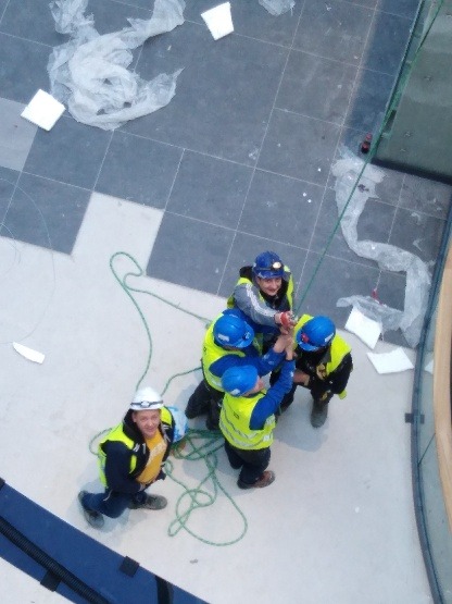 Grupa elektryków w kaskach i kamizelkach odblaskowych, pracujących zespołowo przy instalacji elektrycznej w nowym budynku, widok z góry, z widocznymi kablami i narzędziami.