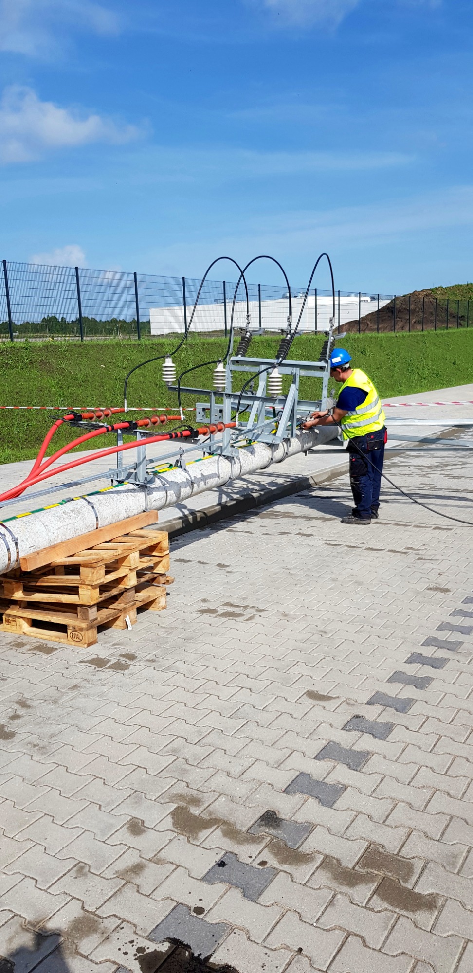 Elektryk w kasku i kamizelce odblaskowej pracuje przy podłączeniu kabli do słupa energetycznego na zewnątrz, widoczne palety podtrzymujące rurę.