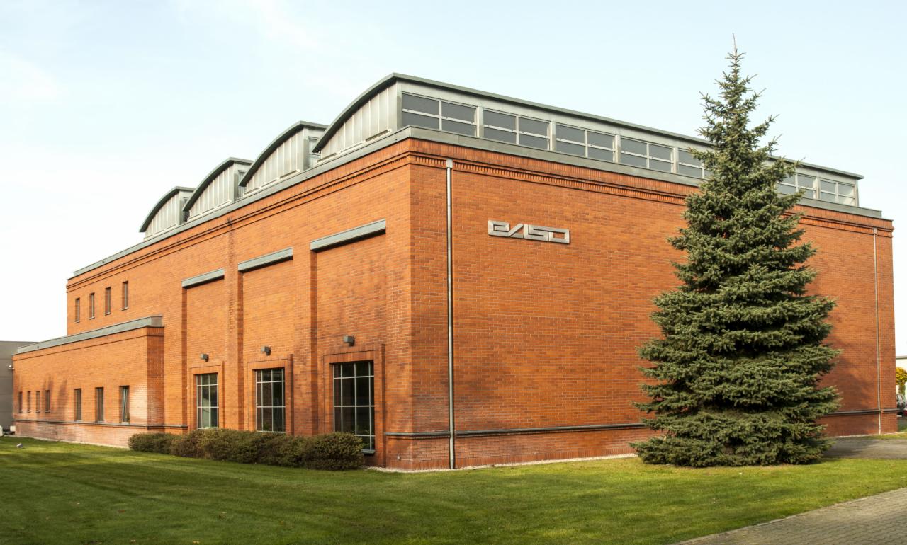 Ceglany budynek z metalowym logo na ścianie, widoczny fragment trawnika i choinka.