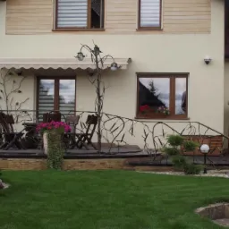 Kuta balustrada z motywem gałęzi przed domem z tarasem i markizą, w tle zadbany trawnik i roślinność.