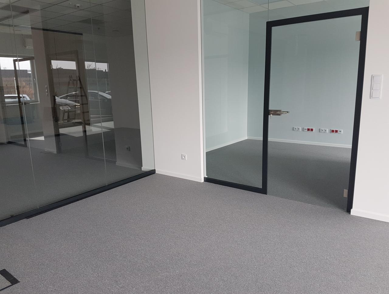 Szara wykładzina dywanowa w nowoczesnym biurze z przeszklonymi ścianami i drzwiami z czarnymi ramami.