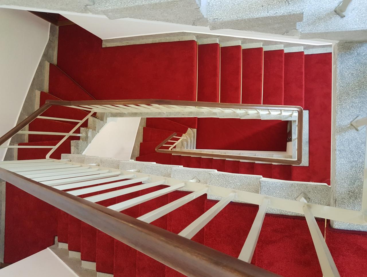Widok z góry na schody pokryte czerwoną wykładziną, białe i drewniane balustrady, szare betonowe stopnie.