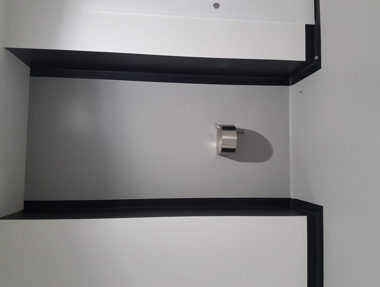 Szara ściana z zamontowanym metalowym uchwytem na papier toaletowy, widoczne czarne listwy przypodłogowe i narożnik ściany.