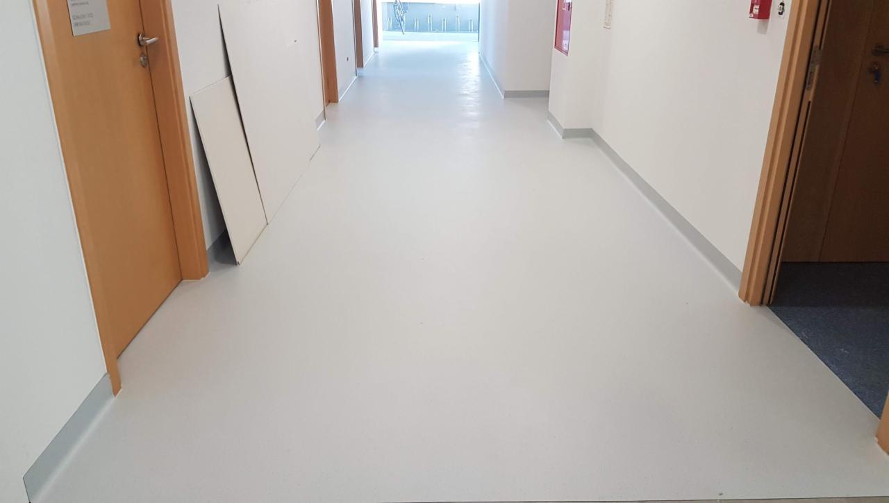 Jasnoszara wykładzina PCV w korytarzu biurowym, widok w kierunku światła dziennego, z widocznymi drzwiami po bokach i panelami na ścianie.