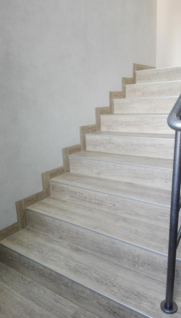 Schody wykończone wykładziną imitującą jasne drewno, z metalowymi listwami na krawędziach stopni i fragmentem metalowej poręczy, przy szarej ścianie.