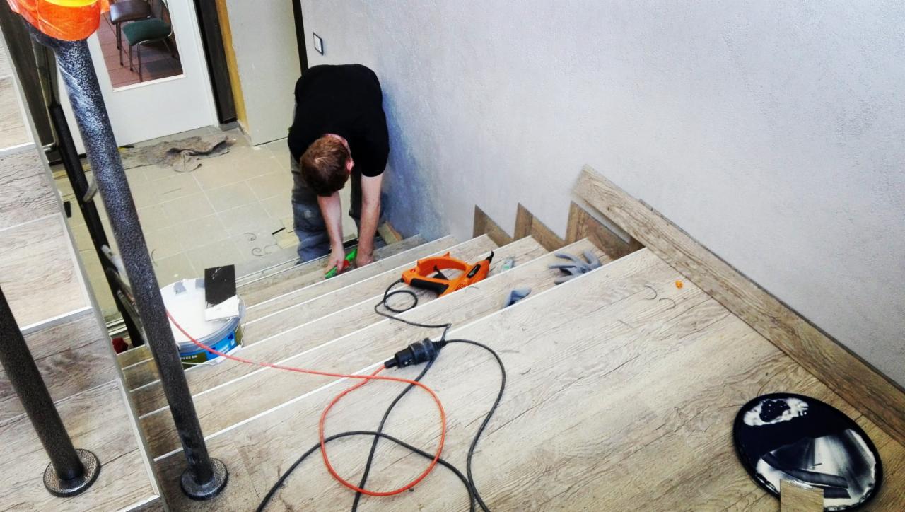 Pracownik w czarnej koszulce przycina wykładzinę na schodach, widoczny zszywacz tapicerski, puszka farby i kable