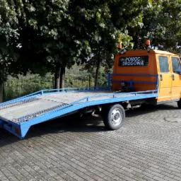 Pomarańczowa laweta z niebieską platformą, zaparkowana na brukowanym placu, z napisem 'Pomoc Drogowa' na tylnej szybie.