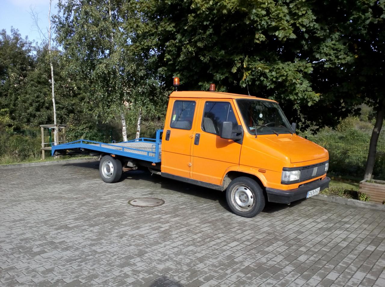 Pomarańczowa laweta z niebieską platformą, zaparkowana na brukowanej powierzchni, otoczona zielenią drzew.