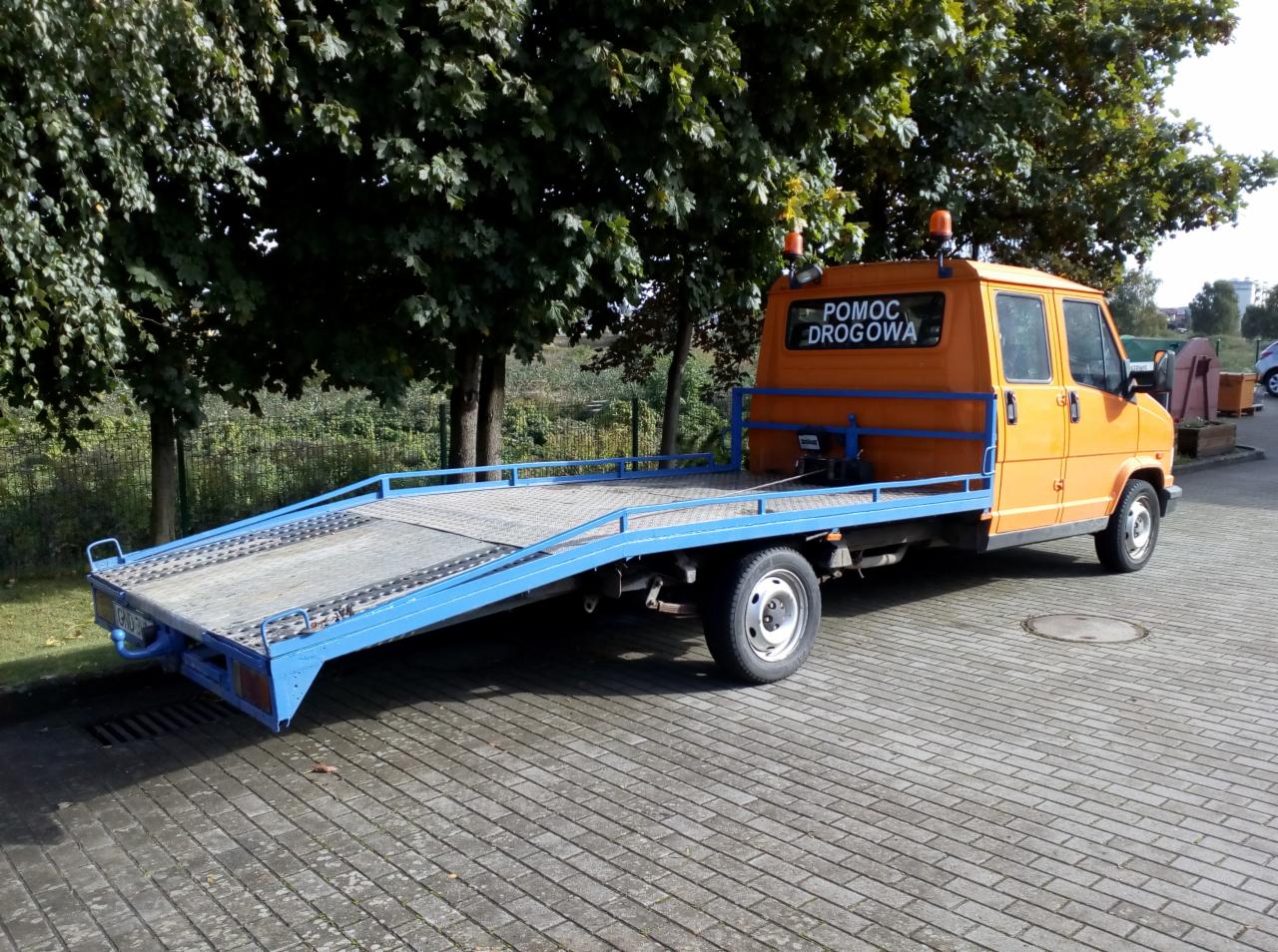 Pomarańczowa laweta z niebieską platformą, zaparkowana na brukowanym placu, z napisem 'Pomoc Drogowa' na tylnej szybie.