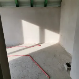 Wnętrze w trakcie prac wykończeniowych: ściany po nałożeniu gładzi, widoczne instalacje elektryczne w czerwonych peszlach na podłodze, surowy betonowy podkład, cień padający na ścianę.