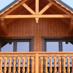 Fragment elewacji drewnianego domu z balkonem, widoczne zdobione balustrady, okna w ciemnych ramach i konstrukcja dachu z dekoracyjnymi elementami wykończeniowymi.