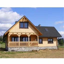 Jasny, dwukondygnacyjny dom z drewna z ciemnym dachem i kamienną podmurówką, z zadaszonym balkonem i ozdobnymi detalami wykończeniowymi, na tle błękitnego nieba.