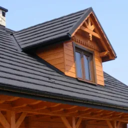 Fragment domu z bali z ciemną dachówką i ozdobnym lukarną, widoczny komin z kamienną obudową.