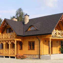 Żółty dom z bali z ciemnym dachem, dwoma kominami z kamienia, drewnianymi zdobieniami i balkonem, otoczony zielenią.