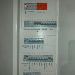 Rozdzielnia elektryczna z opisami obwodów, widoczne wyłączniki różnicowoprądowe i bezpieczniki, zasilanie podpisane dla piekarnika, zmywarki, kuchni, klimatyzacji i rezerwy.