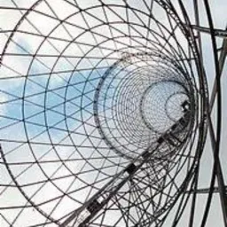 Ażurowa konstrukcja wieży z metalowych prętów widziana z dołu, tworząca tunelowy efekt z niebem w tle. Geometryczny wzór, perspektywa zbieżna.