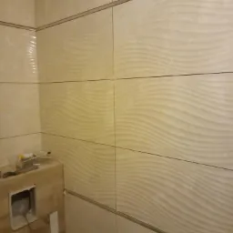 Beżowe płytki ścienne z falistym wzorem w łazience, widoczne narzędzia i akcesoria na blacie, częściowo wykończona ściana.