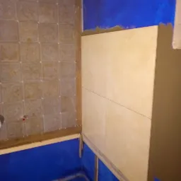 Niedokończona łazienka z częściowo ułożonymi jasnymi płytkami na ścianie i niebieskim zabezpieczeniem podłogi, widoczne listwy montażowe.