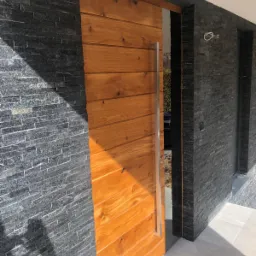 Nowoczesne wejście: drewniana płaszczyzna z metalowym uchwytem sąsiaduje z elewacją z ciemnego kamienia łupkowego.