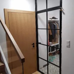 Drewniana ościeżnica z pojedynczym skrzydłem w Katowicach, sąsiadująca z przeszkloną ścianką działową z czarnymi szprosami, za którą widoczna garderoba i wzorzysta podłoga.