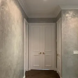 Korytarz z podwójnymi białymi drzwiami i dekoracyjną szarą ścianą o teksturze przypominającej marmur, wykończony białymi listwami przysufitowymi