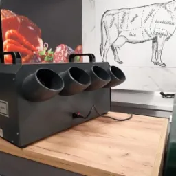 Czarna, przemysłowa suszarka do wędlin z czterema wylotami powietrza, stojąca na drewnianym blacie w zakładzie przetwórstwa mięsnego, z widoczną grafiką podziału tuszy wołowej na ścianie w tle.