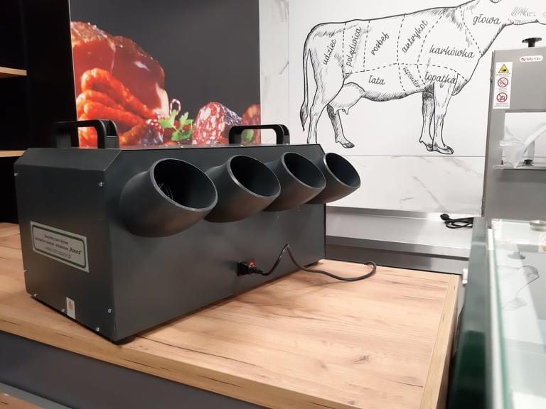 Czarna, przemysłowa suszarka do wędlin z czterema wylotami powietrza, stojąca na drewnianym blacie w zakładzie przetwórstwa mięsnego, z widoczną grafiką podziału tuszy wołowej na ścianie w tle.