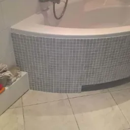 Narożna wanna obudowana mozaiką w szarych odcieniach, jasne płytki na podłodze i ścianach, półka z ręcznikami obok wanny.