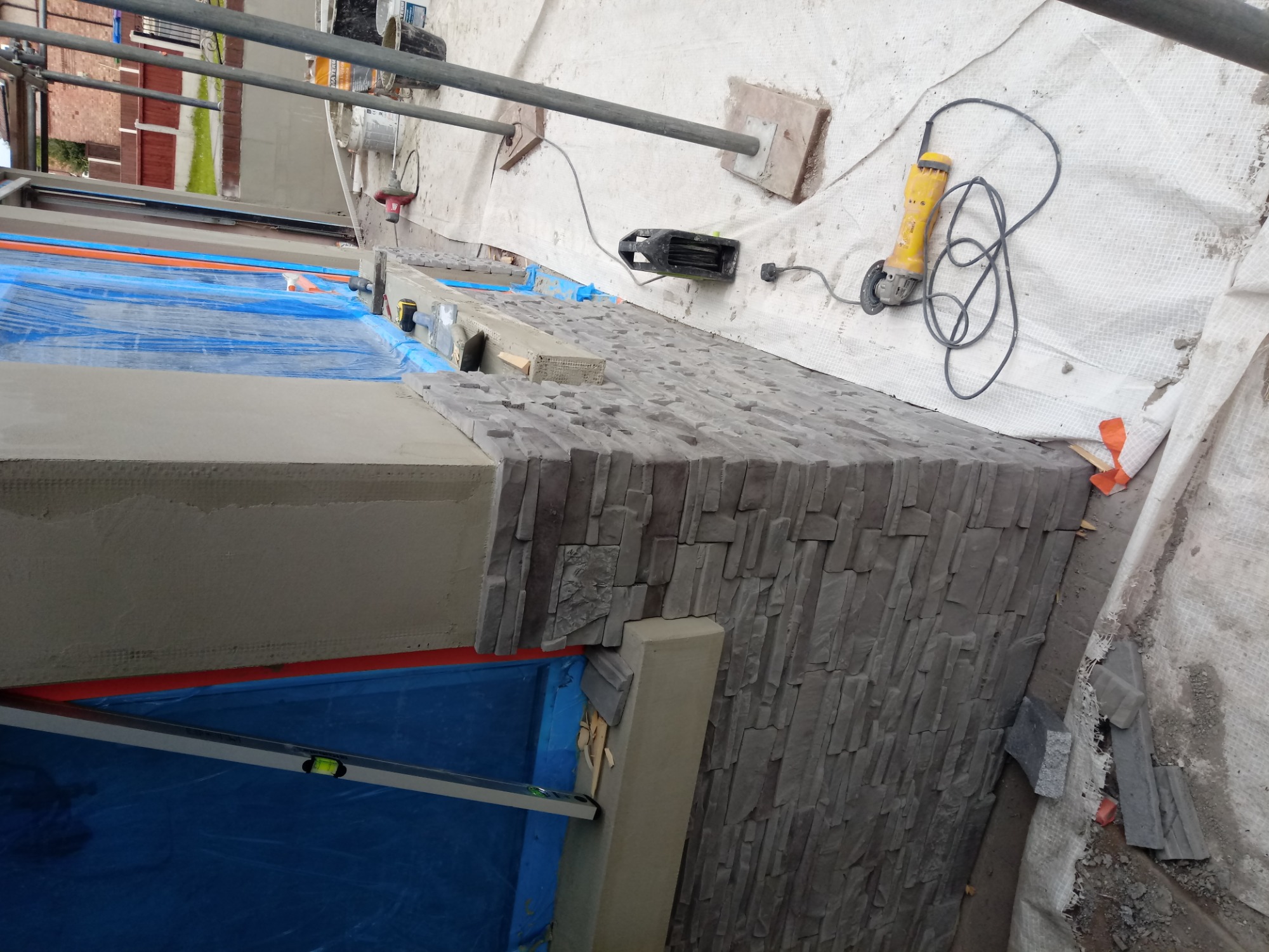 Montaż dekoracyjnej okładziny kamiennej na ścianie, widoczne narzędzia, poziomica i szlifierka kątowa, zabezpieczone folią podłoże.