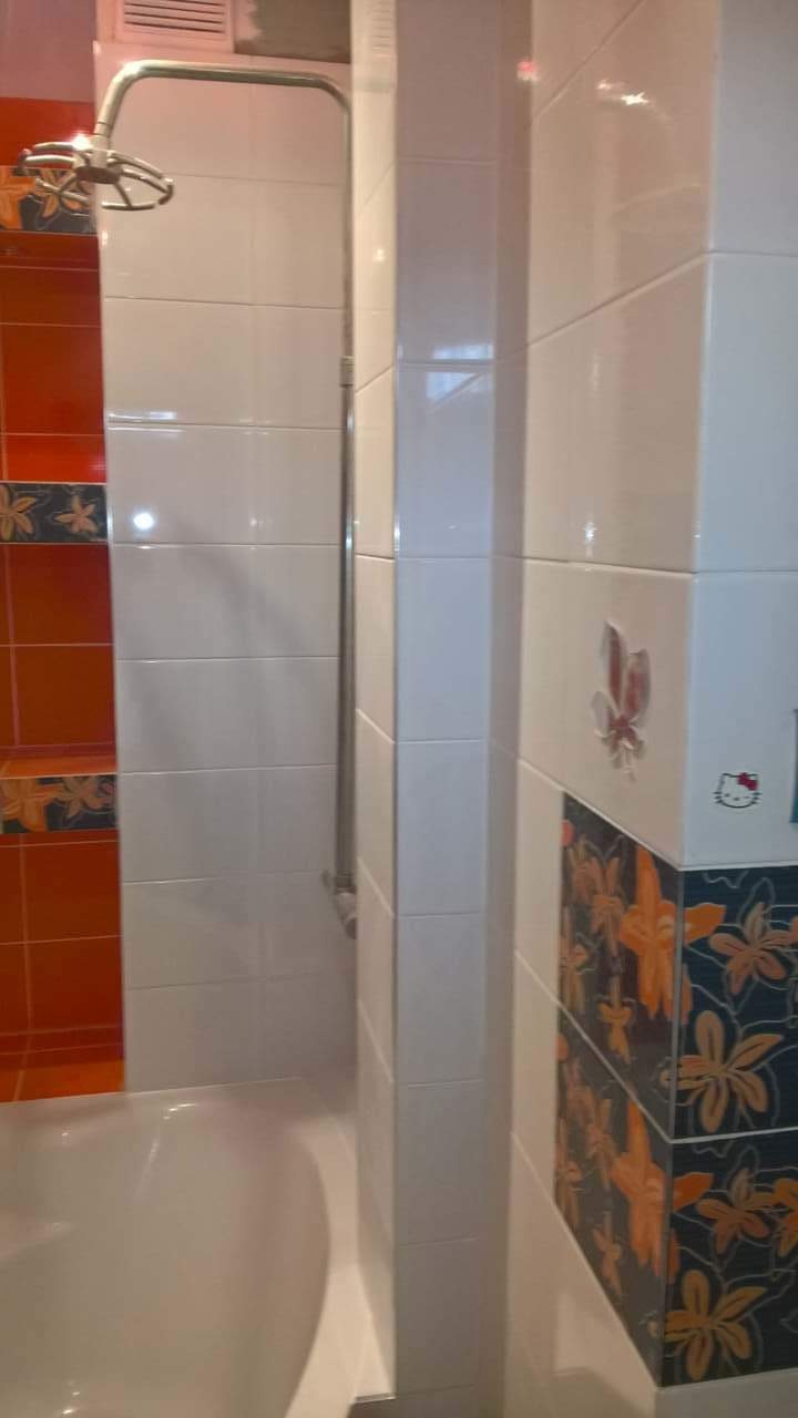 Wykończona łazienka z wanną, białe płytki z motywem kwiatowym i pomarańczowymi akcentami, bateria prysznicowa na ścianie, dekoracyjna naklejka Hello Kitty na kafelkach.