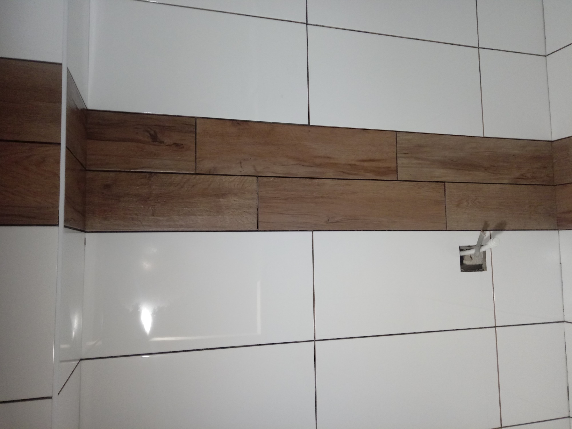 Ściana łazienki wyłożona białymi płytkami z poziomym pasem płytek imitujących drewno, widoczny otwór na instalację.