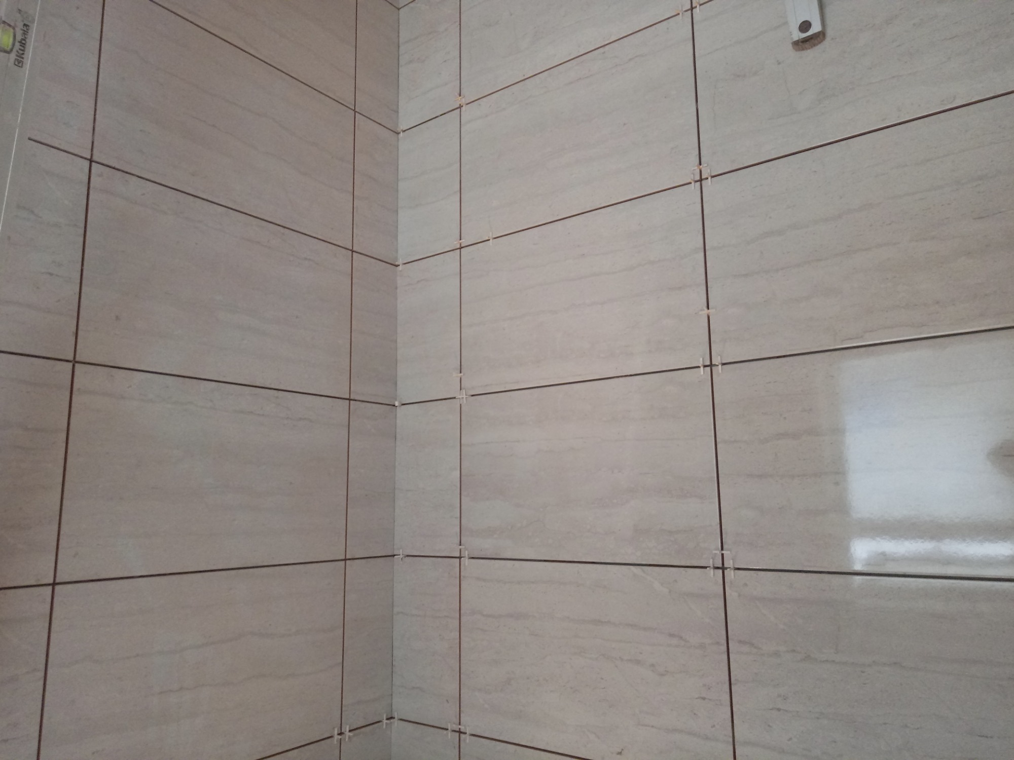 Świeżo ułożone, beżowe płytki imitujące kamień na ścianach w rogu łazienki, z widocznymi krzyżykami dystansowymi i odblaskiem światła na jednej z płytek.