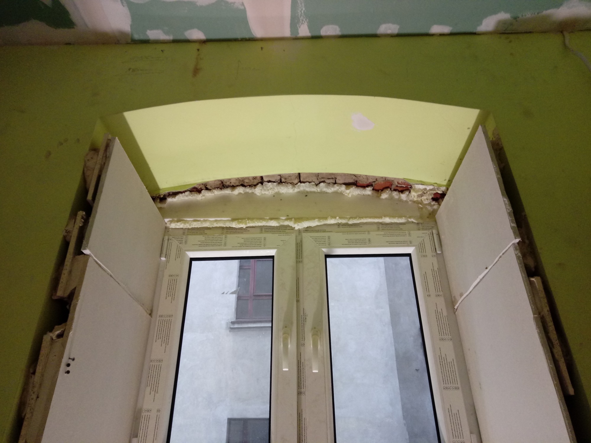 Montaż okna z łukowatym nadprożem, widoczne płyty gipsowe, pianka montażowa i fragment muru pod zieloną ścianą.
