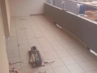 Balkon w trakcie układania płytek podłogowych, widoczna przecinarka do glazury i fragmenty płytek, surowy stan wykończenia, widok na zewnątrz.