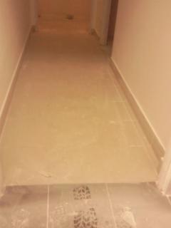 Ułożone jasne płytki podłogowe w wąskim korytarzu, widoczne ślady butów na dolnych płytkach przy wejściu.