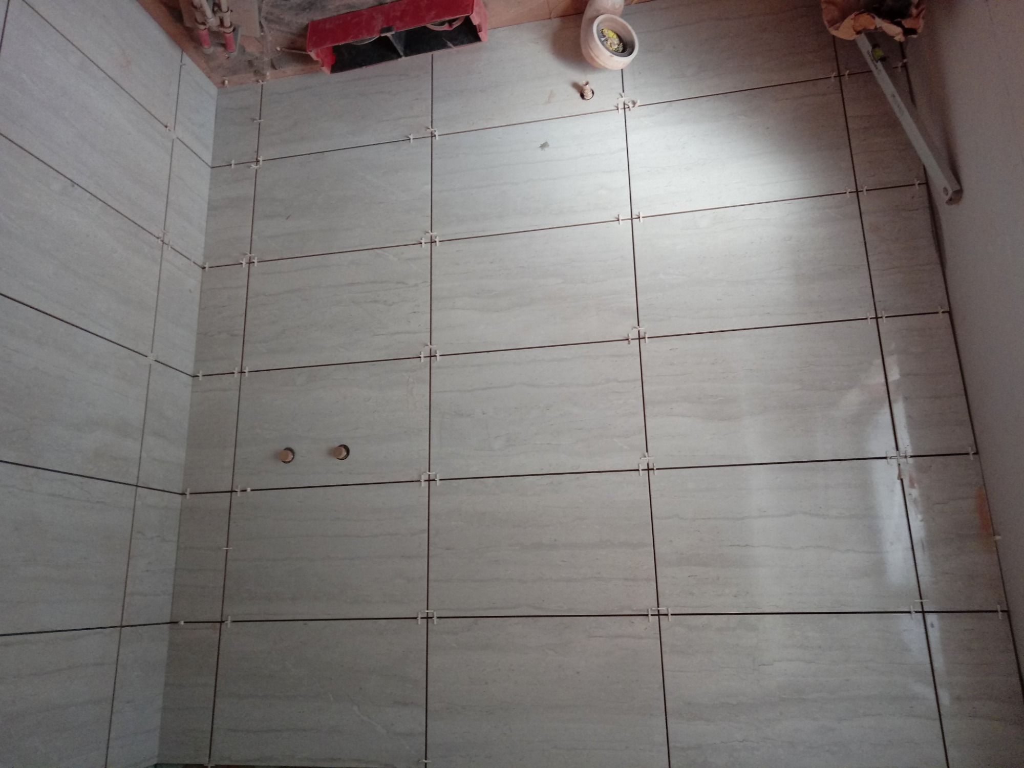 Ściana łazienki w trakcie układania płytek z widocznymi krzyżykami dystansowymi, w tle narzędzia i instalacja hydrauliczna.