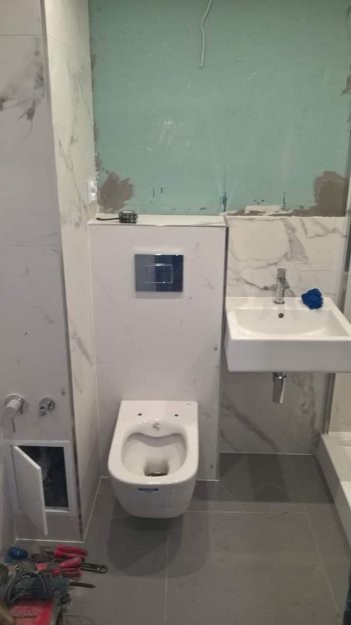 Wnętrze łazienki w trakcie prac wykończeniowych: zamontowana toaleta podwieszana z chromowanym przyciskiem, umywalka z baterią, płytki imitujące marmur na ścianach, szare płytki podłogowe, widoczne...